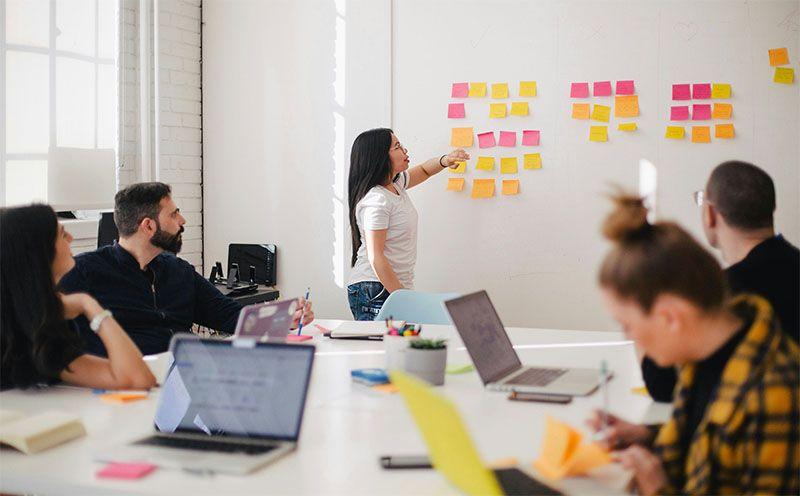 En kvinne skriver på en vegg med fargerike post-it-lapper mens kolleger ser på i et moderne kontormiljø.