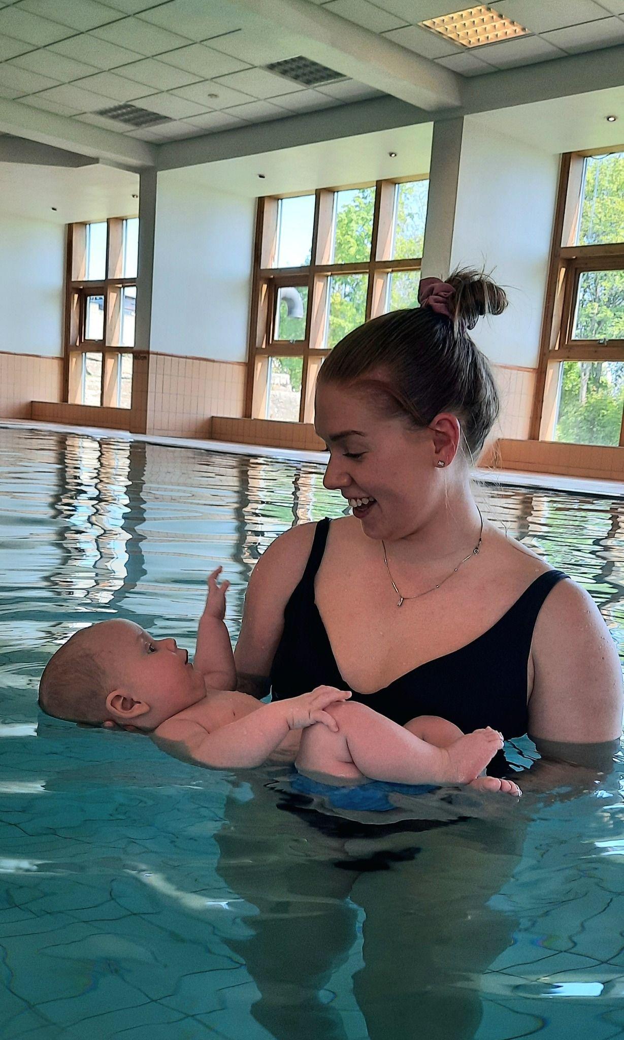 Ung kvinne holder en baby i vannet i et svømmebasseng med store vinduer.
