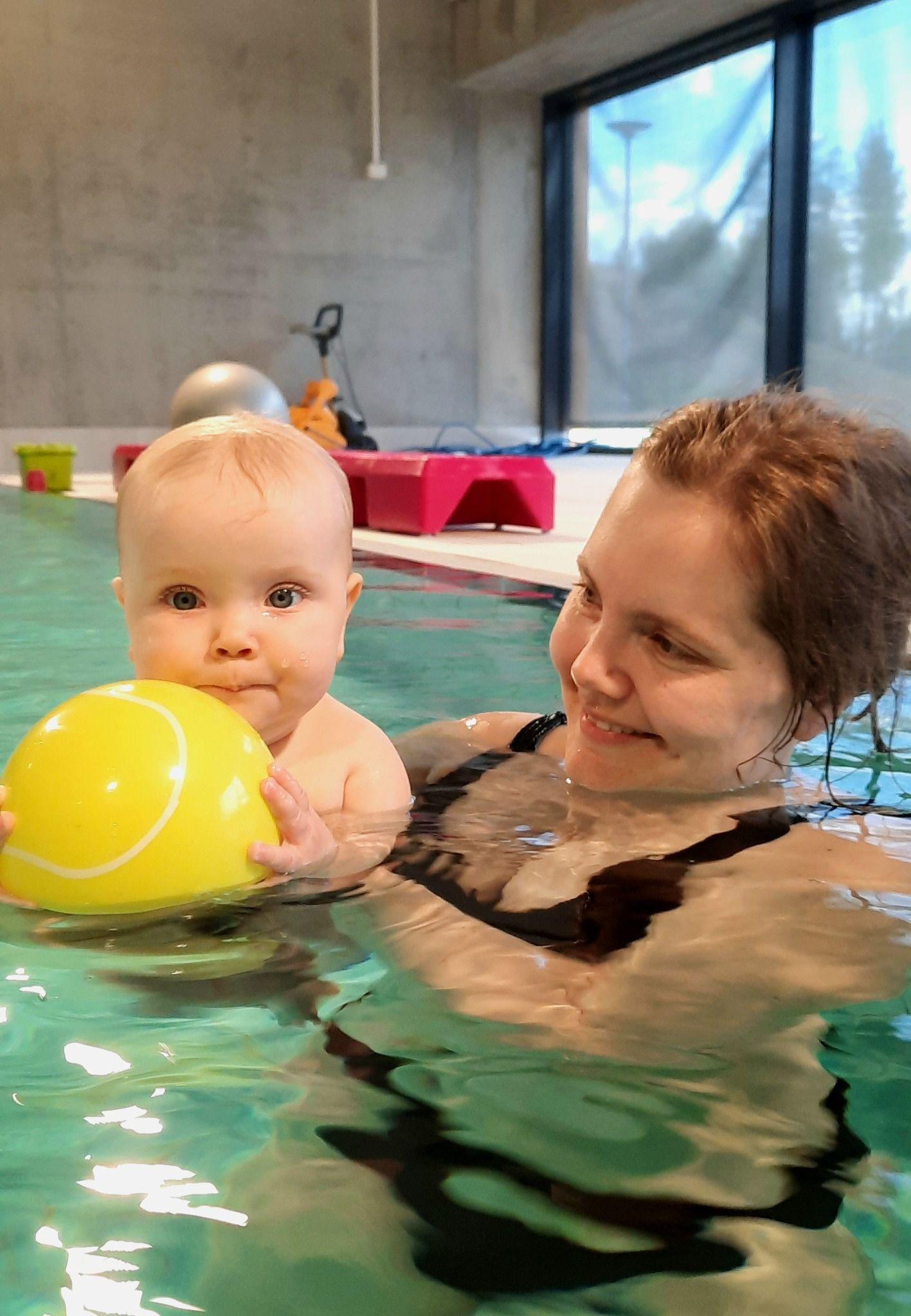 En baby holder en gul ball mens hun svømmer med en voksen kvinne i et svømmebasseng.