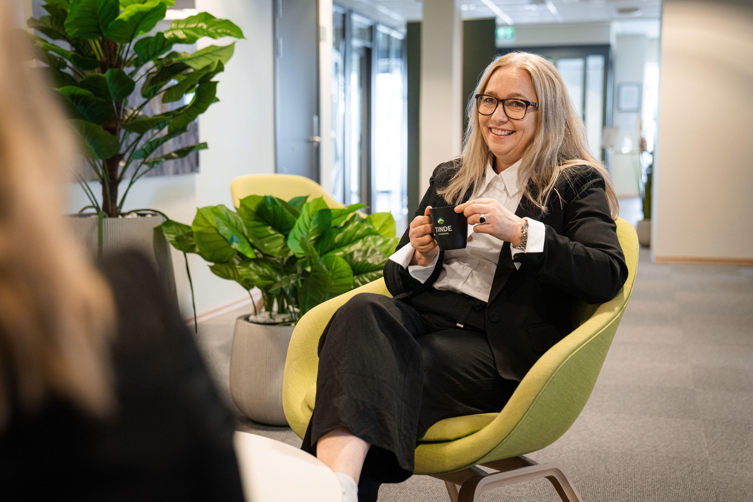 En kvinne med langt hår sitter i en grønn stol og holder en kopp, omgitt av planter i et moderne kontor.