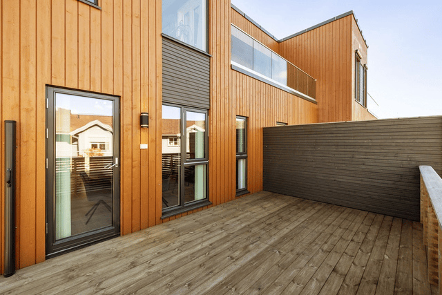 Moderne hus med trepanel og terrasse, med store vinduer og en romslig uteplass i solfylt vær.