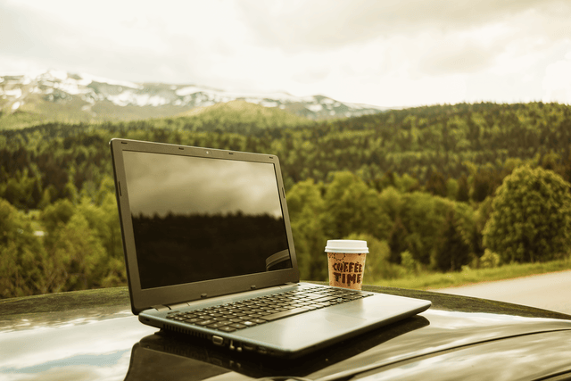 En bærbar datamaskin og en kaffekopp står på en bil, med fjell og skog i bakgrunnen.