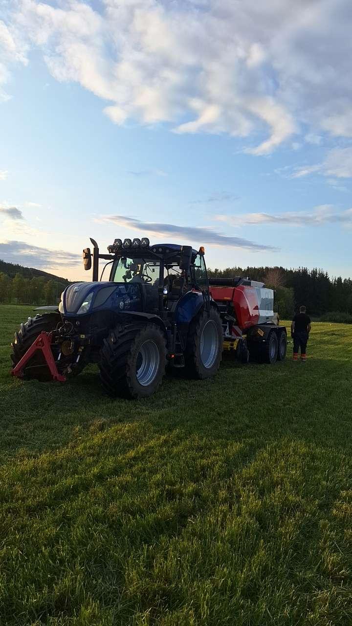 En svart traktor med tilhenger står på en grønn eng under en blå himmel med skyer. To personer jobber i nærheten.