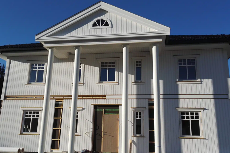 Moderne, hvitt hus med søyler og store vinduer under bygging i solfylt vær.
