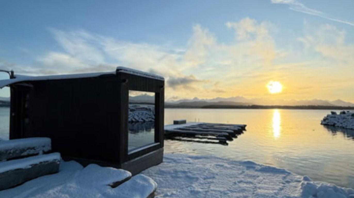 En vinterlig solnedgang over en rolig innsjø med en moderne hytte dekket av snø.