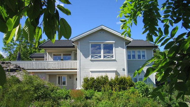 Moderne, lysegrått hus med balkong, omringet av grønne planter og trær under en klar blå himmel.