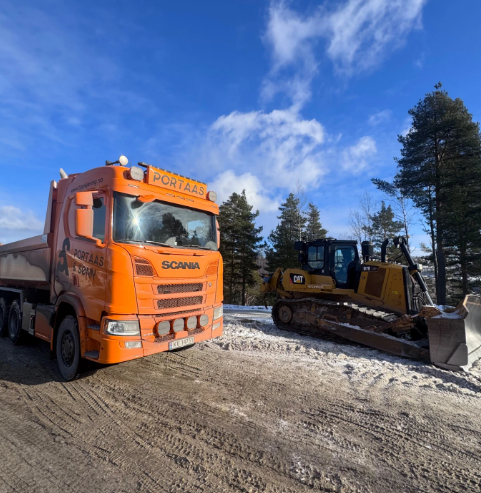 En oransje Scania lastebil står ved siden av en gravemaskin på en snødekt vei med trær i bakgrunnen.