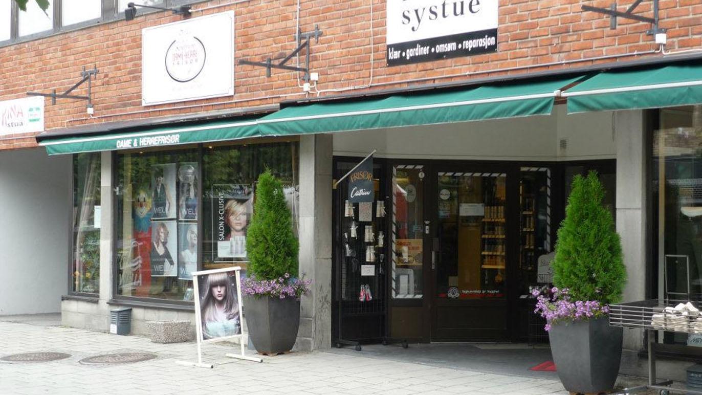 Inngangen til en butikk med grønne markiser, blomsterkasser og reklameplakater på veggen.
