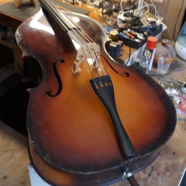 En gammel kontrabass med en slitt overflate, plassert på et arbeidsbord med verktøy og utstyr i bakgrunnen.