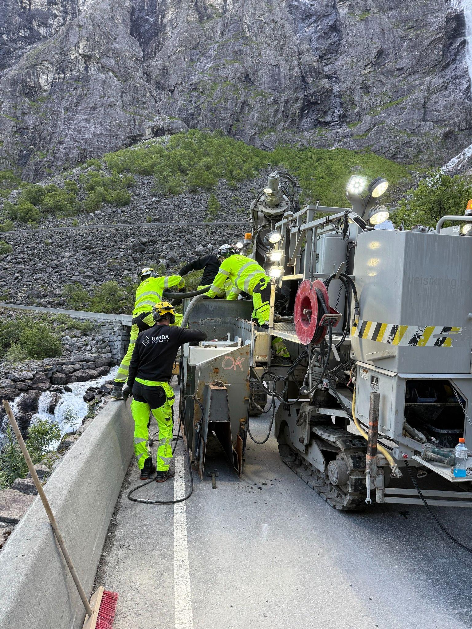 Arbeidere i neon gule klær jobber med en stor maskin langs en vei i fjellandskap.