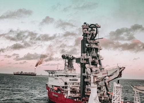 Et stort offshore skip med kran og rørledninger, med en annen båt i bakgrunnen under en fargerik himmel.