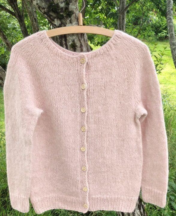En lys rosa strikket cardigan med knapper, hengende på en kleshenger i en grønn hage.