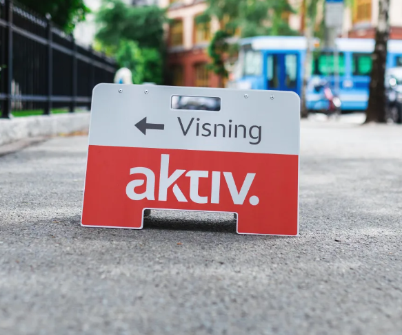 Skilt med teksten "Visning" og "aktiv" som står på fortauet, med en bygate i bakgrunnen.