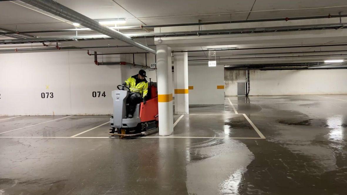 En person kjører en gulvvaskemaskin i en parkeringskjeller med vått gulv og nummererte parkeringsplasser.