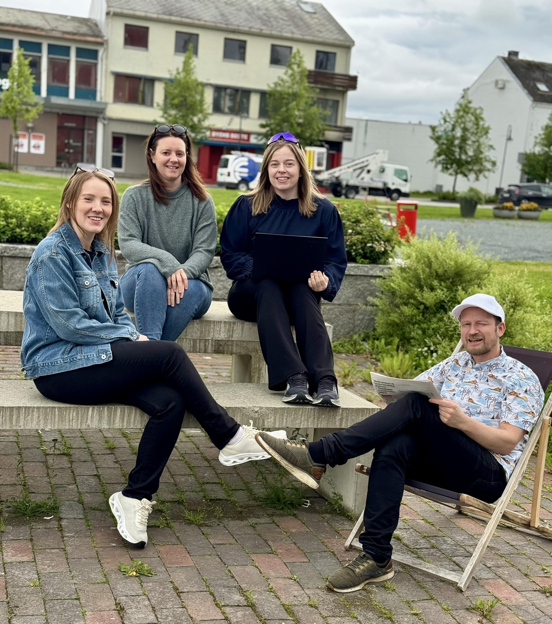 Fire personer sitter på en benk i en bypark, smilende og engasjert i samtale. Grønne trær og bygninger i bakgrunnen.