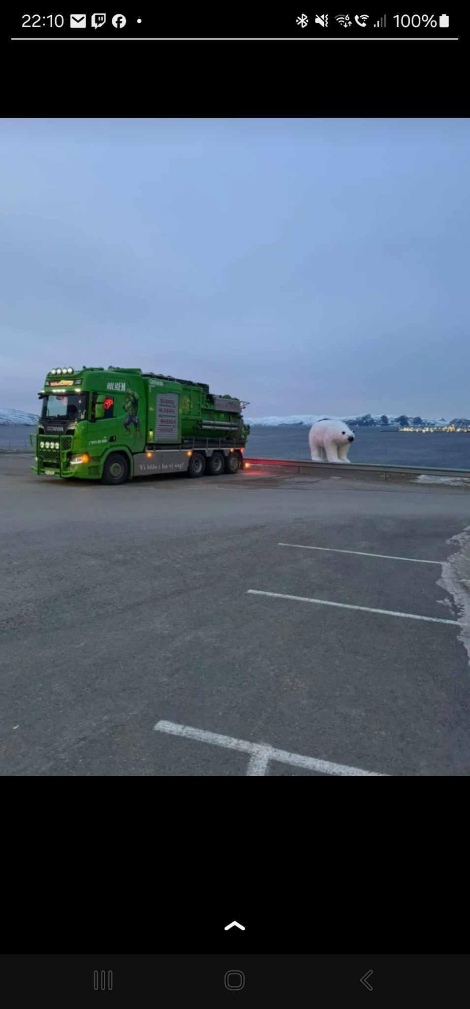 Grønn lastebil parkerer ved vannkanten med en stor hvit gjenstand i bakgrunnen.