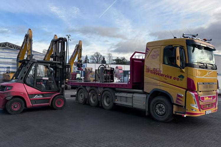Lastebil med flatbed og gaffeltruck på en byggeplass, med gravemaskiner i bakgrunnen.