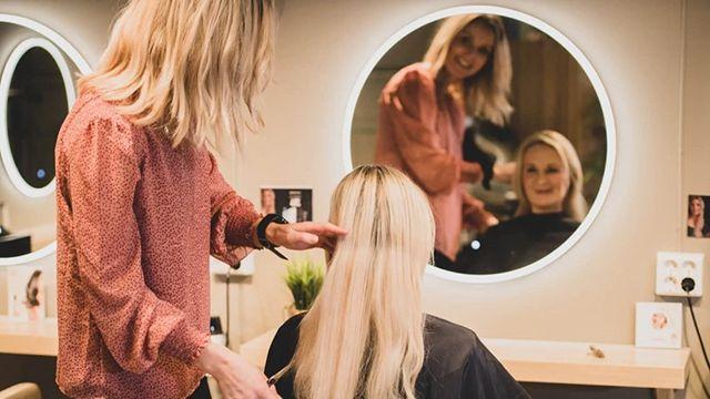 En frisør styler langt hår på en kunde foran et speil i en moderne frisørsalong.