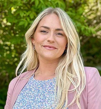 Ung kvinne med langt, lyst hår, iført en rosa blazer og blomstret topp, står foran en grønn bakgrunn.