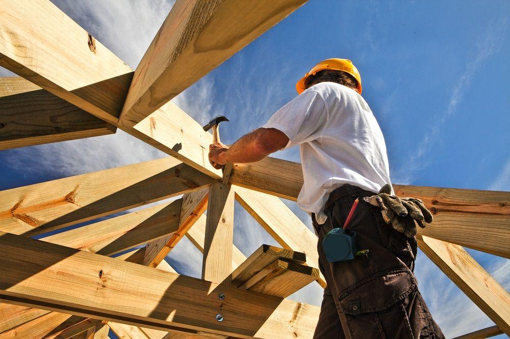 En håndverker som bruker hammer på takstoler under bygging av et hus, med blå himmel i bakgrunnen.