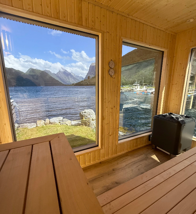 Utsikt fra en hytte med store vinduer mot fjorden og fjellene, med en trebenk i forgrunnen.