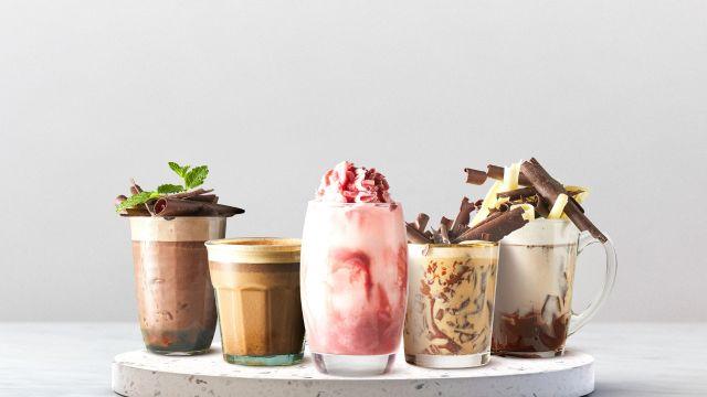 Fem forskjellige milkshakes i glass, inkludert sjokolade, jordbær og karamell, dekorert med sjokoladebiter og mynte.