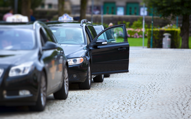 Svart taxi med åpen dør står parkert på en brosteinsbelagt gate i en bypark.