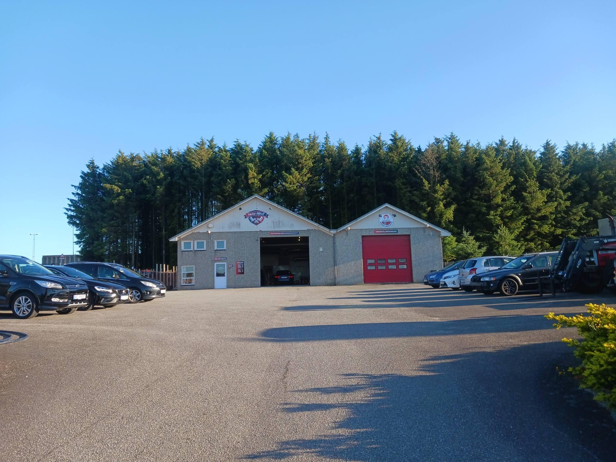 En bilforhandler med flere biler parkert foran en grå bygning, omgitt av trær under en klar blå himmel.