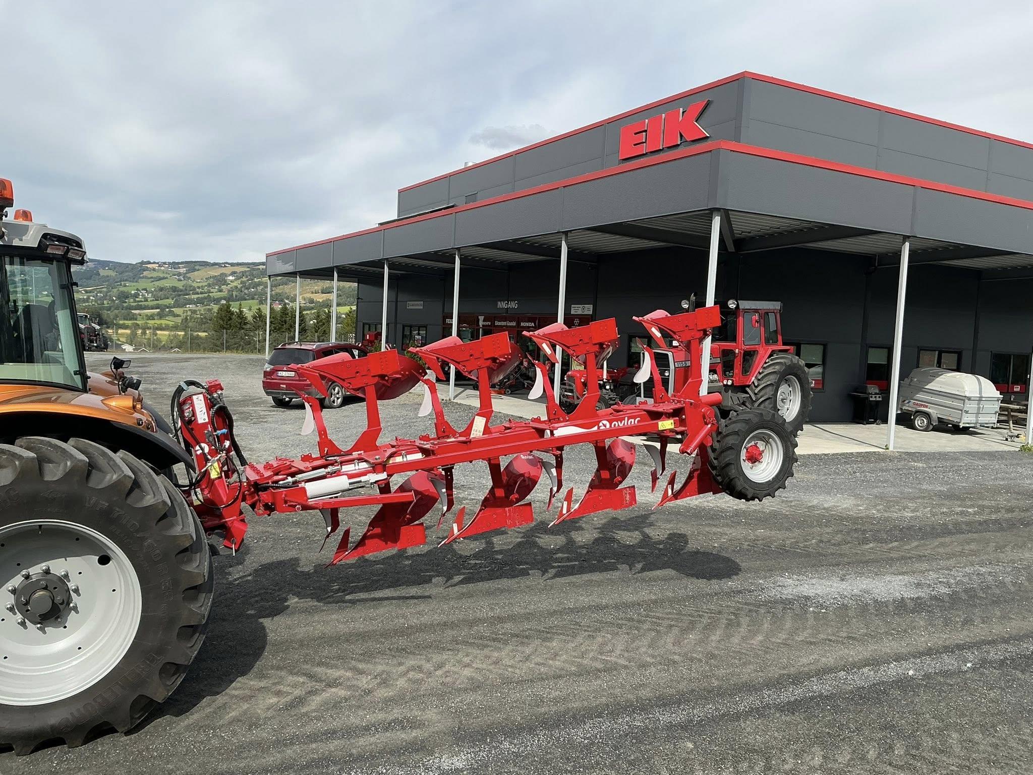 En rød plog festet til en traktor foran en moderne bygning med EIK-logo.