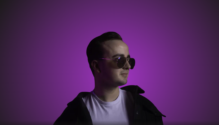 Ung mann med solbriller og svart jakke, poserer mot en lilla bakgrunn.