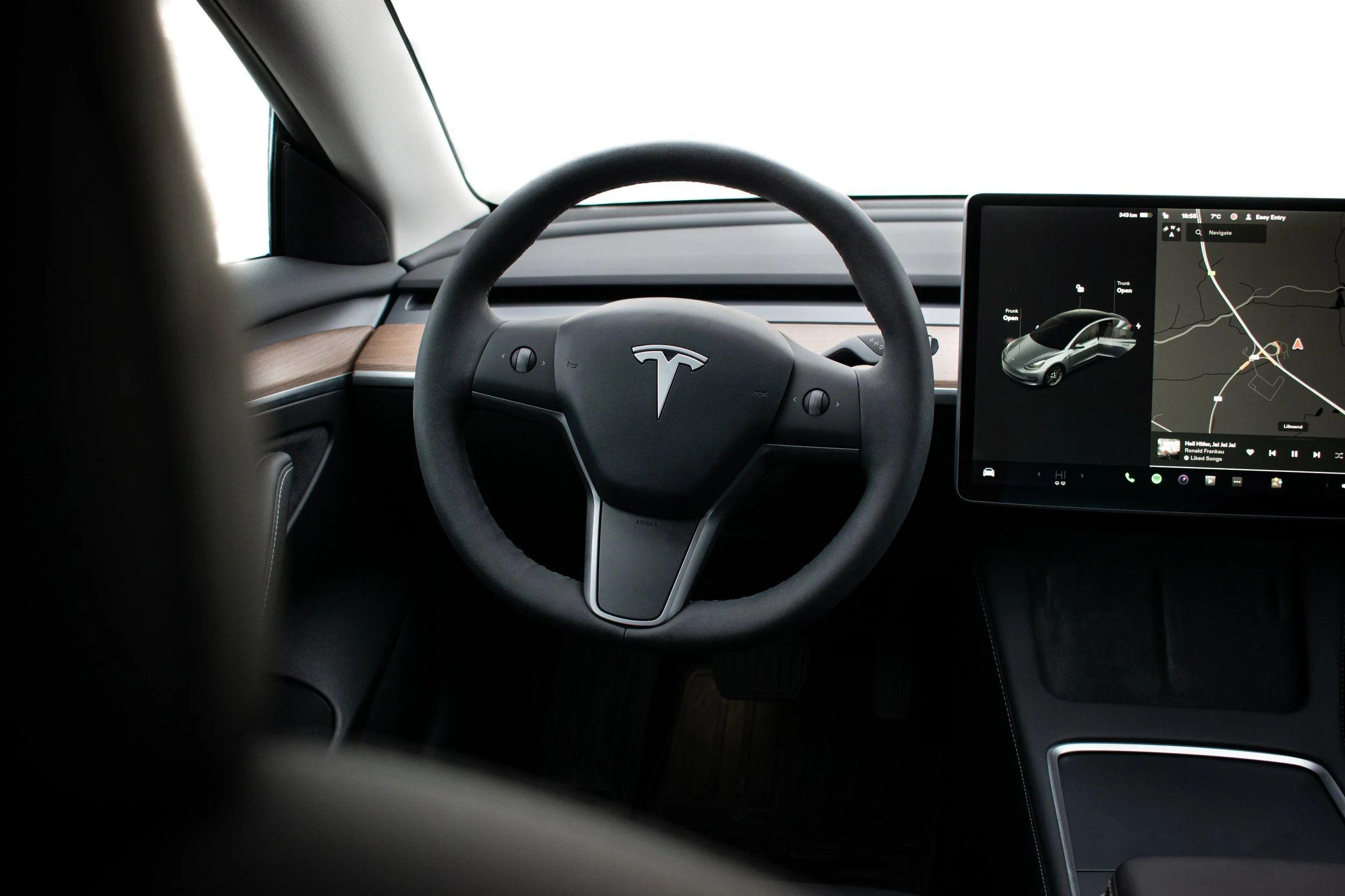 Interiør av en Tesla med ratt og berøringsskjerm som viser navigasjon og bilens kontroller.