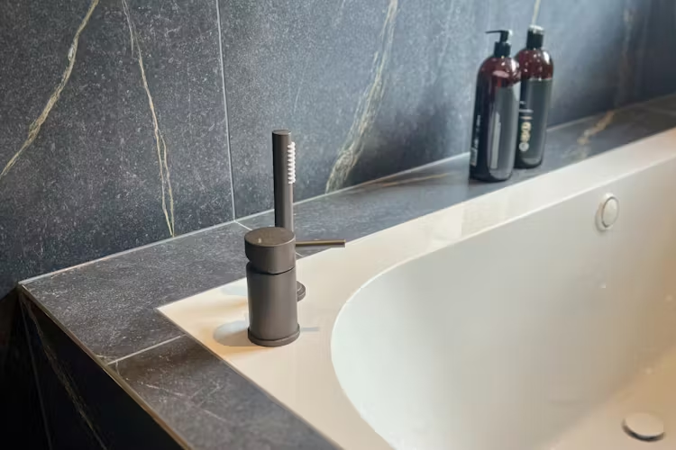 Moderne badekar med svart marmoroverflate og stilren kran, samt flasker med badeprodukter i bakgrunnen.