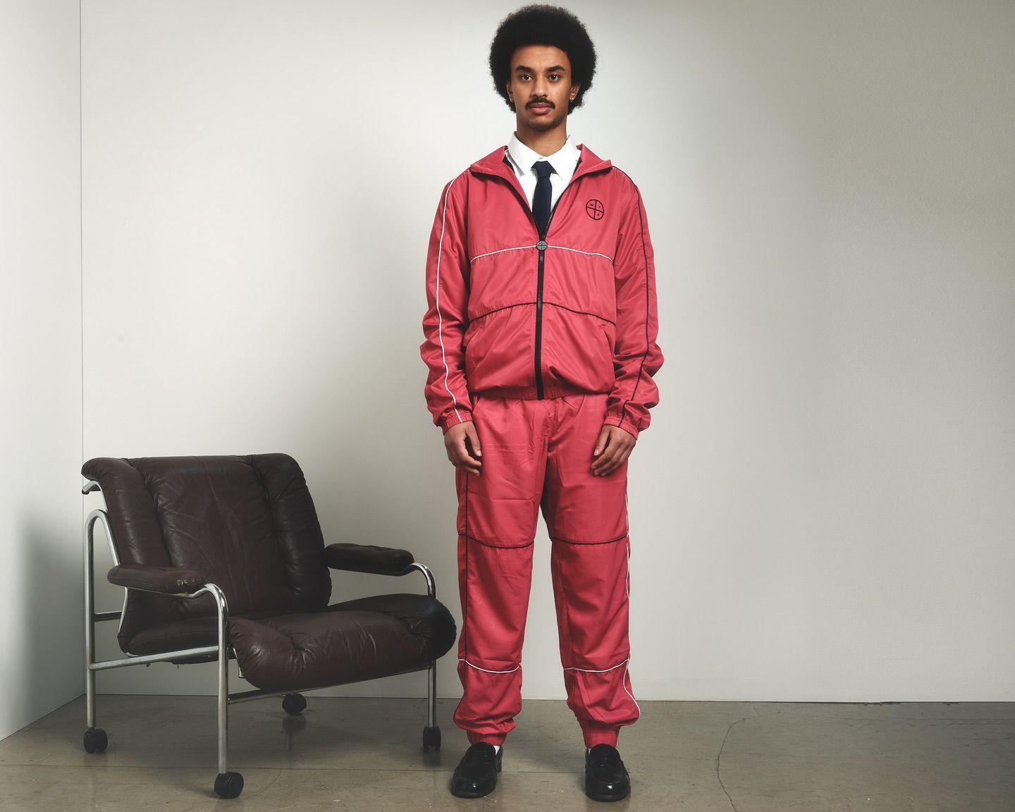 Mann i rød treningsdrakt med afrofrisyre, står foran en brun stol i et minimalistisk rom.