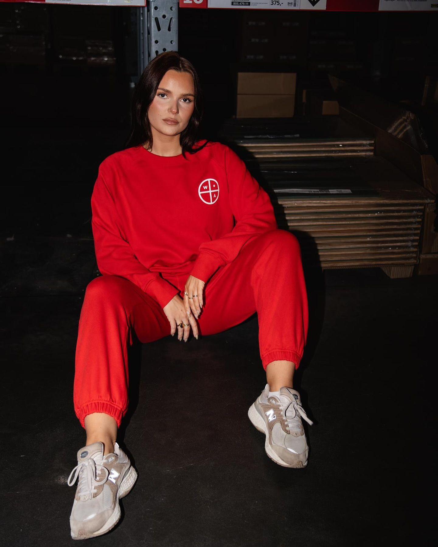 Kvinne i rød joggedress sitter på gulvet med grå sneakers i et lagerlokale.