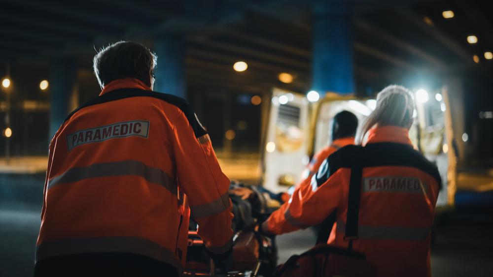 Ambulansepersonell i oransje uniformer som frakter en båre under en bro om natten.
