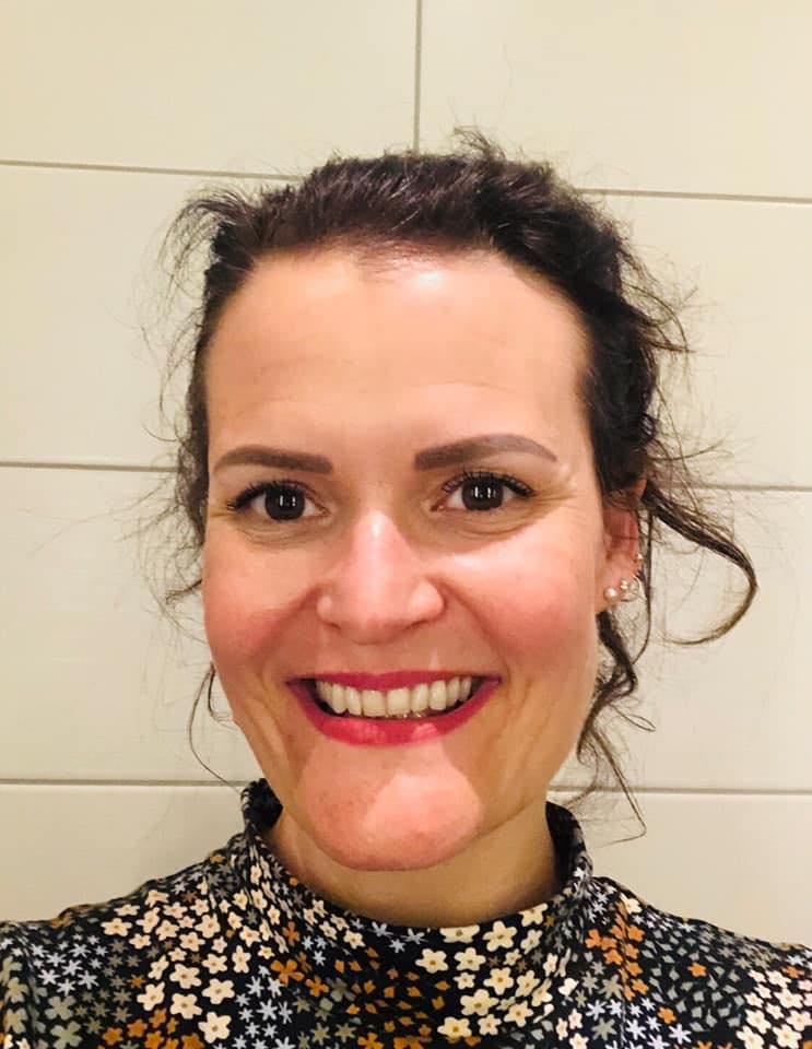 En kvinne med mørkt hår og smilende ansikt, iført en blomstret topp, står foran en lys bakgrunn.
