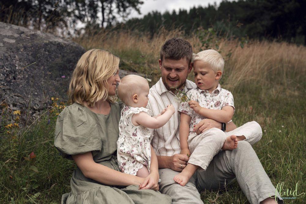 Familie med to små barn sitter på gresset, omringet av natur, og smiler mens de leker sammen.