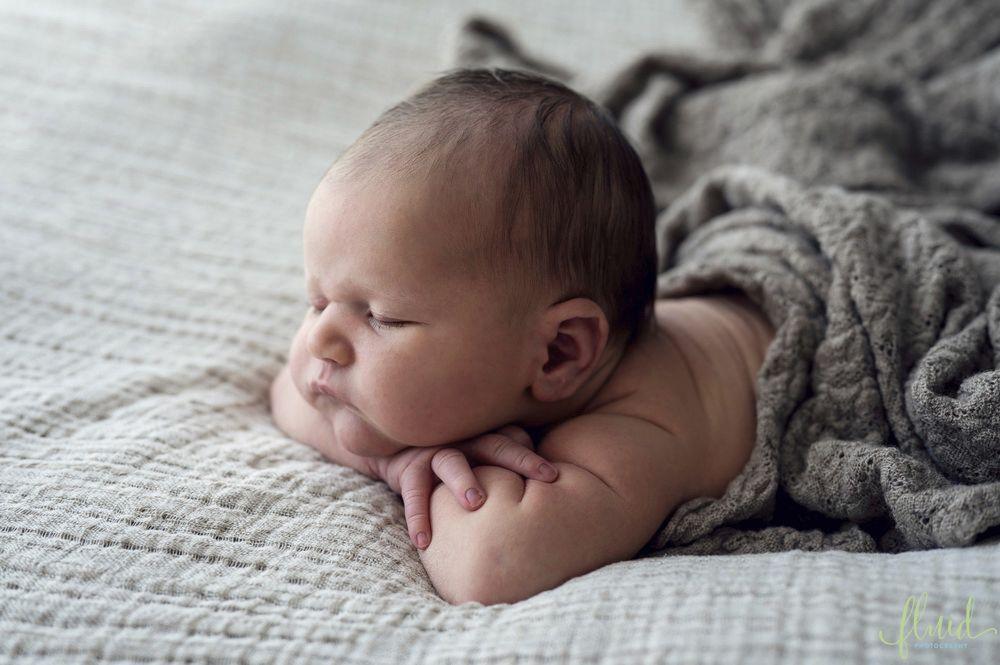 Nyfødt baby ligger på en seng med et grått teppe, med hodet hvilt på armene og et rolig uttrykk.