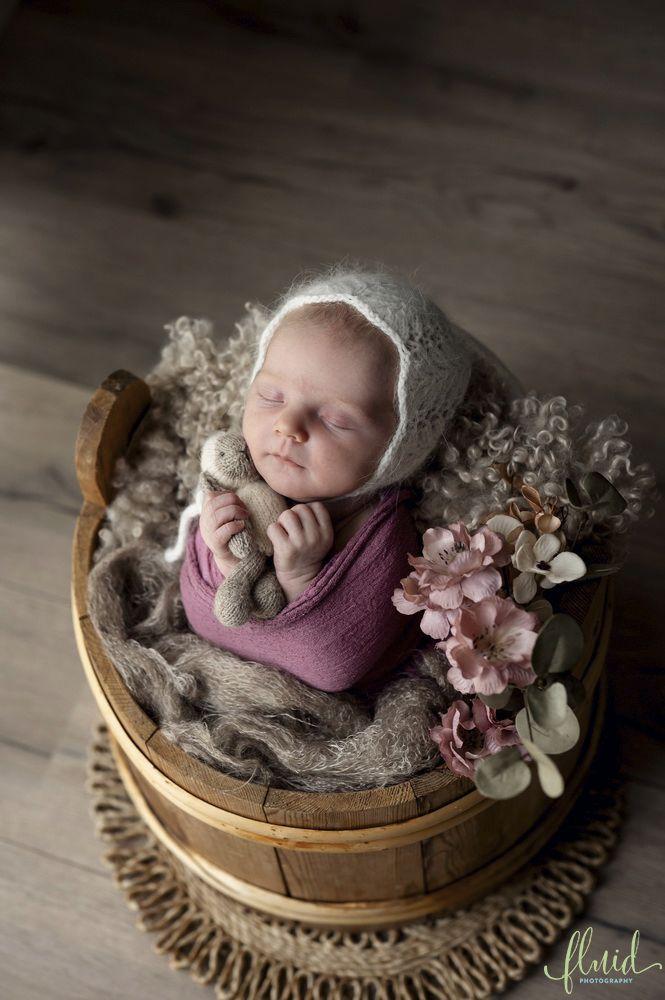 Nyfødt baby i en kurv med blomster, iført en hvit lue og lilla genser, sover fredelig på en myk pute.