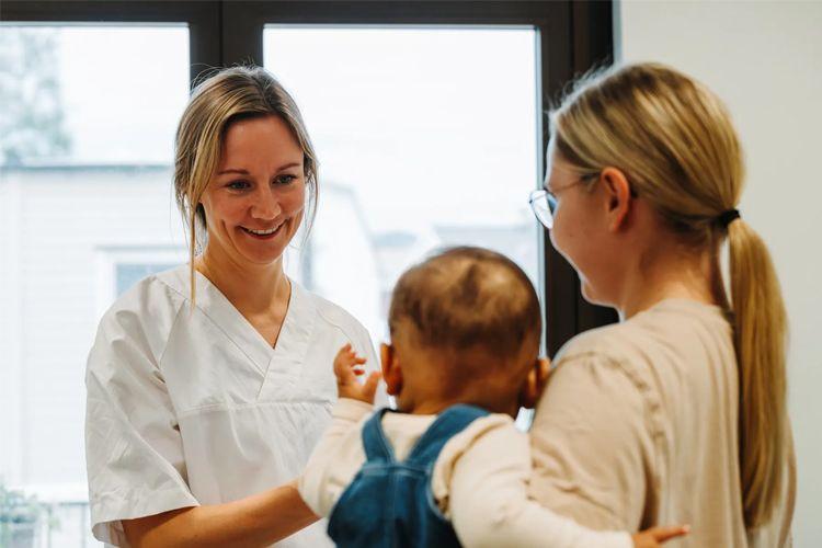 En smilende helsearbeider snakker med en mor som holder et lite barn i armene.