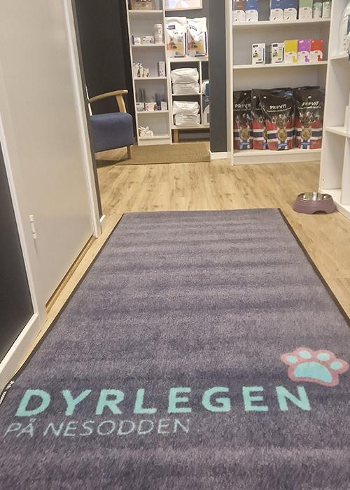 En inngang til en dyreklinikk med en teppe som har teksten "DYRLEGEN PÅ NESODDEN" og hyller med dyreprodukter.