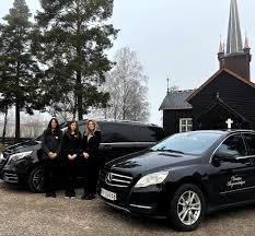 Tre kvinner i svarte klær står foran en svart kirke med to begravelsesbiler parkert.