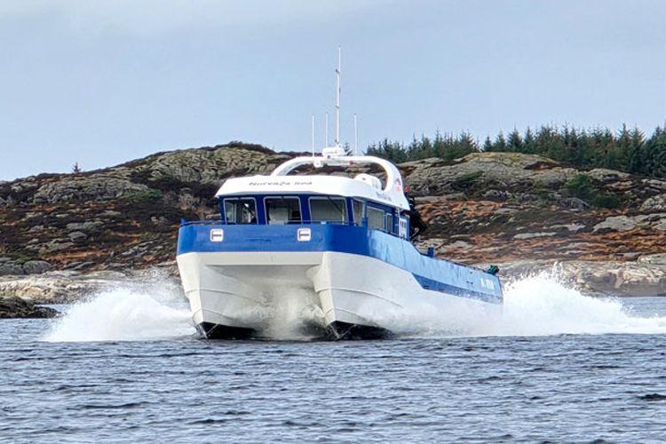 En hurtigbåt med blå og hvit farge som kjører gjennom bølgene i en skjærgård. Grønn vegetasjon i bakgrunnen.