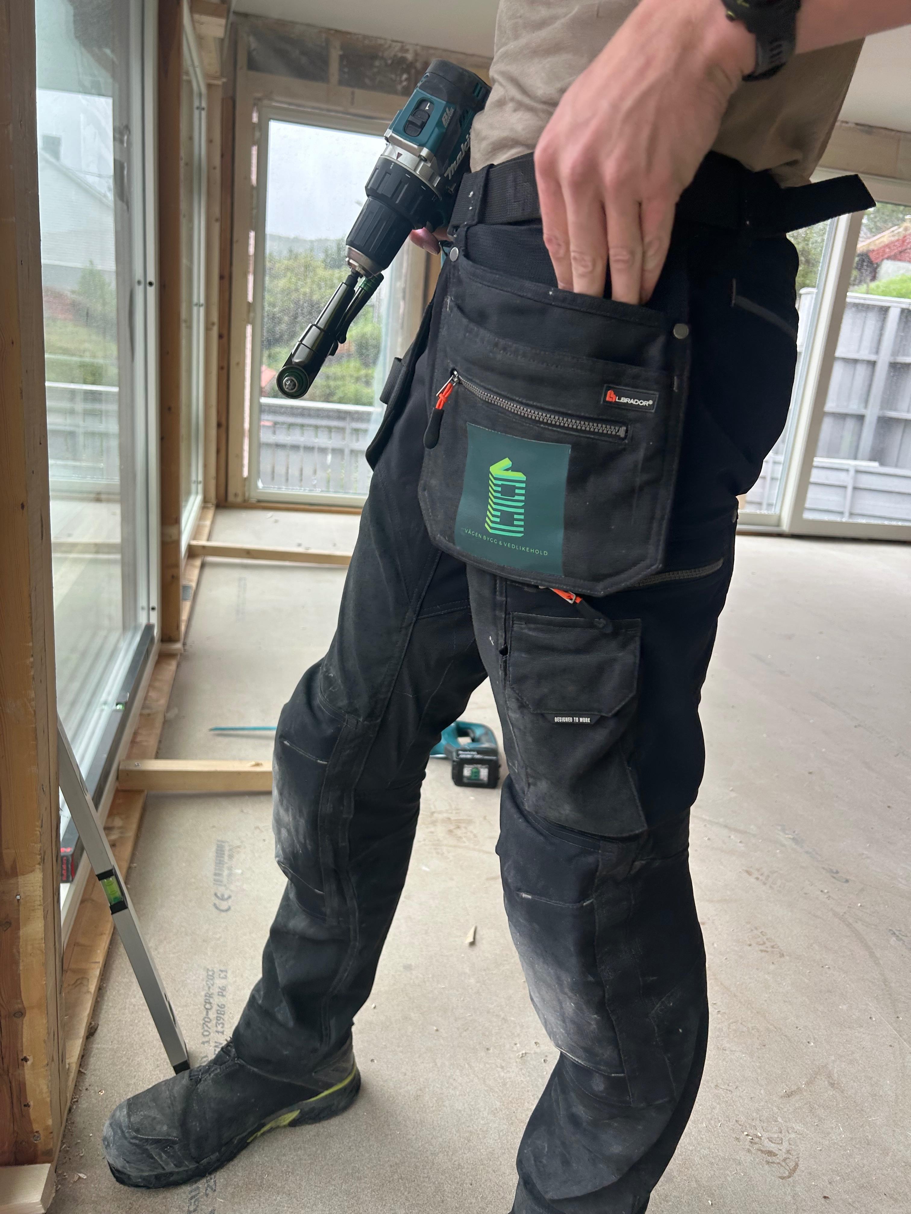 En håndverker i svarte arbeidsbukser står inne i et rom med verktøy i lommen og en drill i hånden.