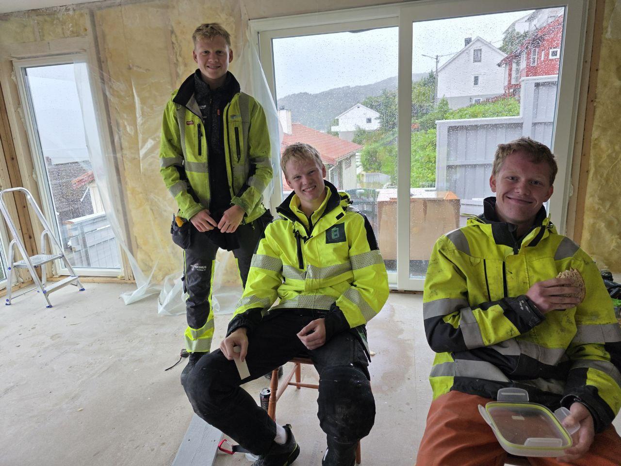 Tre unge menn i arbeidsklær sitter inne i et rom under oppussing, med vinduer og bygninger synlig i bakgrunnen.