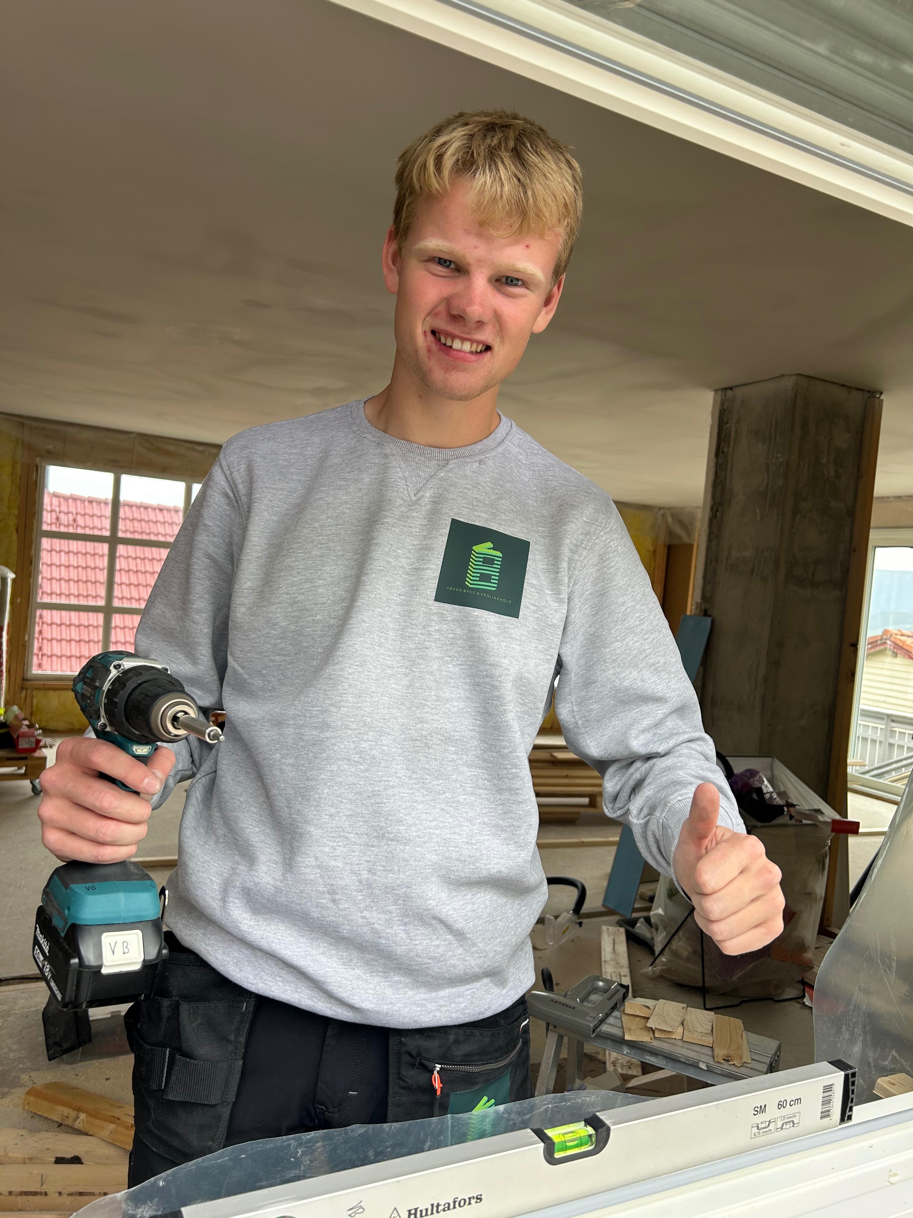 En ung mann med drill står i en byggeplass og gir tommel opp, iført grå genser og arbeidsbukser.