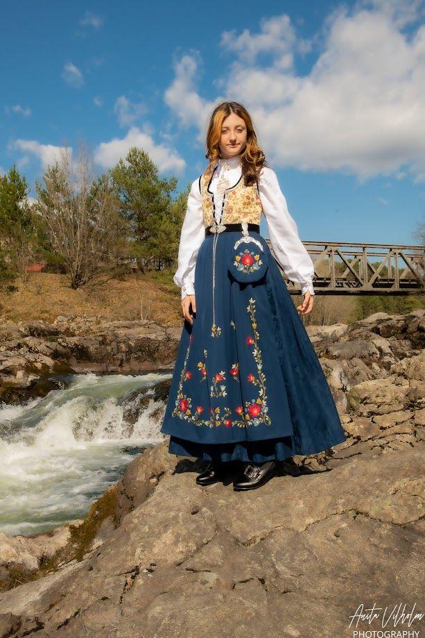Kvinne i tradisjonell bunad med broderier, står ved en foss med blå himmel og trær i bakgrunnen.