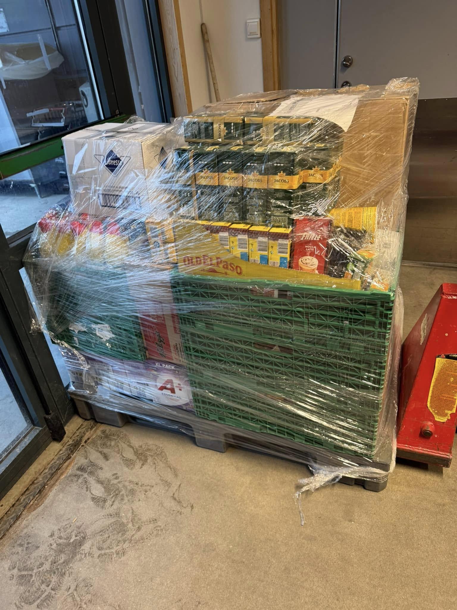 Pall med innpakket varer, inkludert kasser med mat og drikke, står i en lagerlokale.