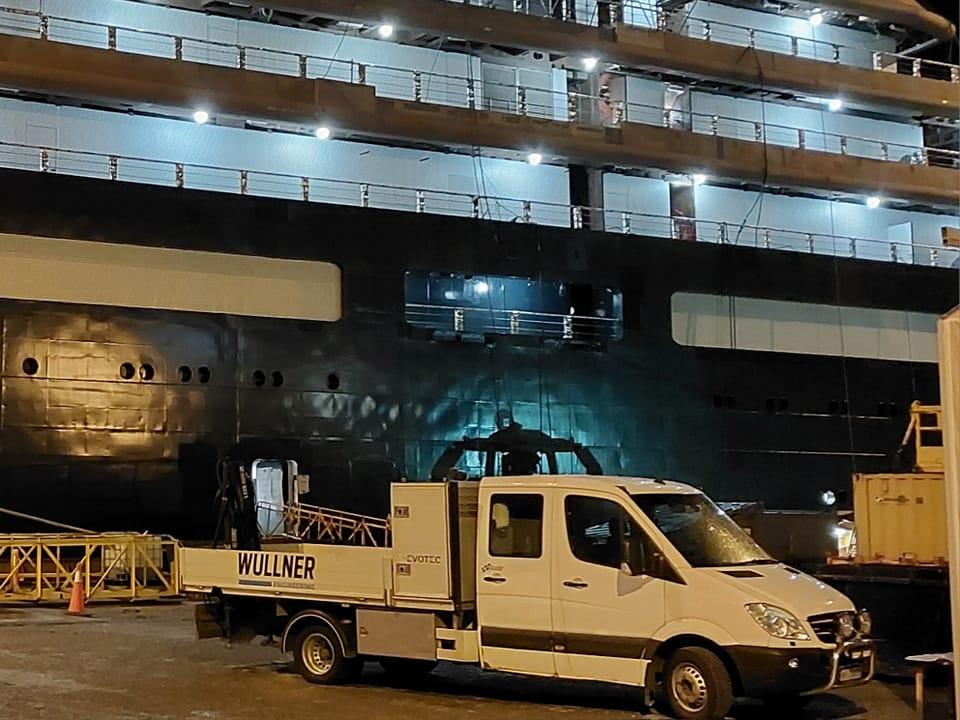 En hvit servicebil står foran et stort svart skip ved kai, med lys som lyser opp området.