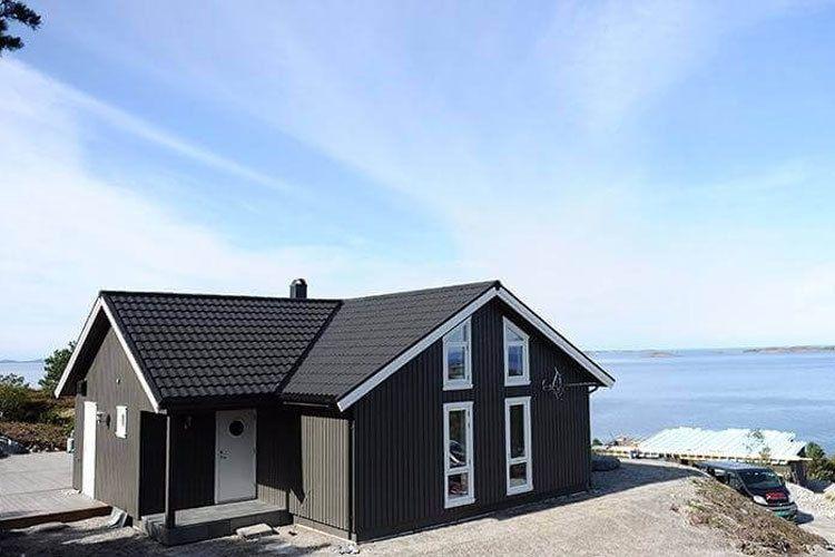 Moderne enebolig med svart kledning og skråtak, beliggende ved sjøen med utsikt over vannet.